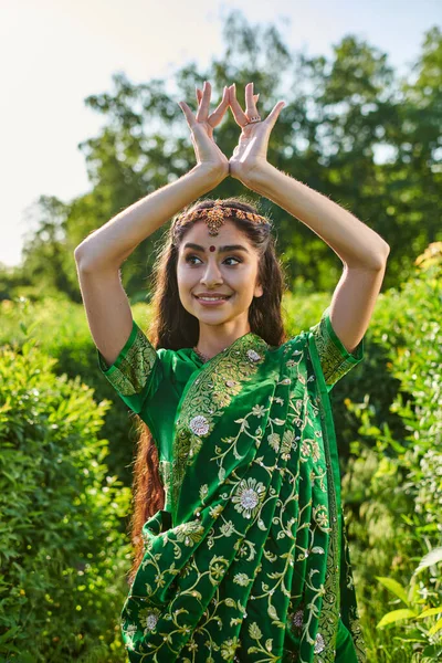 Jovem mulher indiana alegre em sari verde e bindi gestos perto de plantas no parque — Fotografia de Stock