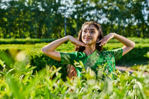 Безтурботний молода індійська жінка в зеленому сарі позує біля кущів в парку влітку — стокове фото