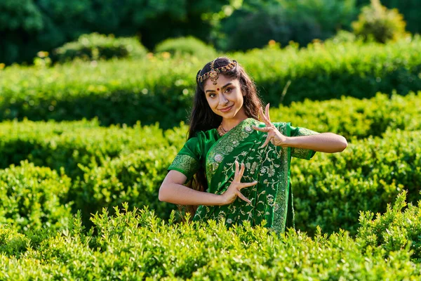 Jeune femme indienne à la mode en sari vert souriant et posant près des buissons dans le parc en été — Photo de stock