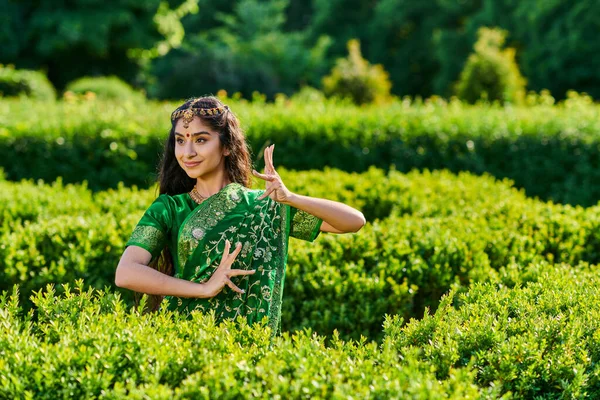 Mujer india joven positiva y elegante en sari verde gesticulando cerca de las plantas en el parque - foto de stock