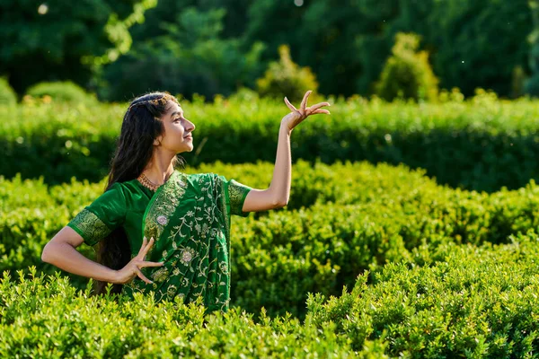 Вид сбоку улыбающейся и стильной индийской женщины в зеленом сари, позирующей возле растений в парке — стоковое фото