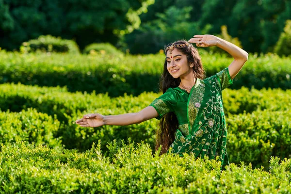 Позитивная молодая индийская женщина в зеленом сари и бинди позирует возле зеленых растений в летнем парке — стоковое фото