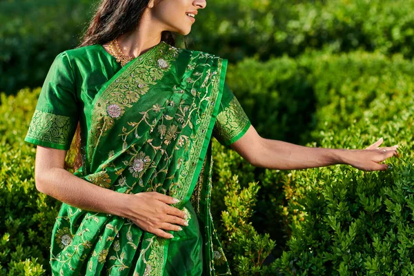 Cropped view of stylish smiling young woman in green sari with pattern touching plants in park — Stock Photo