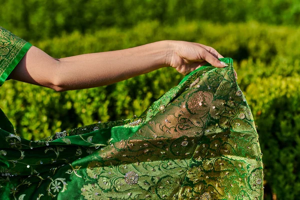 Обрезанный вид молодой женщины, касающейся современного зеленого сари с узором рядом с растениями в парке — стоковое фото