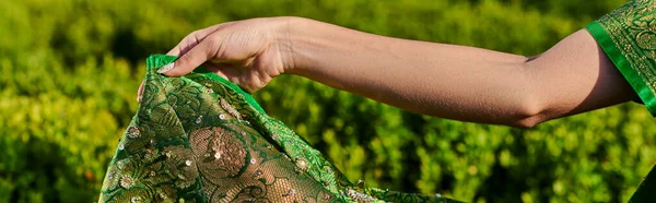 Teilansicht einer jungen Frau, die einen modernen grünen Sari mit Muster in der Nähe von Pflanzen im Park berührt, Banner — Stockfoto