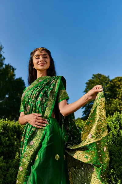 Alegre y moderna mujer india joven tocando sari con patrón cerca de plantas verdes en el parque al aire libre - foto de stock