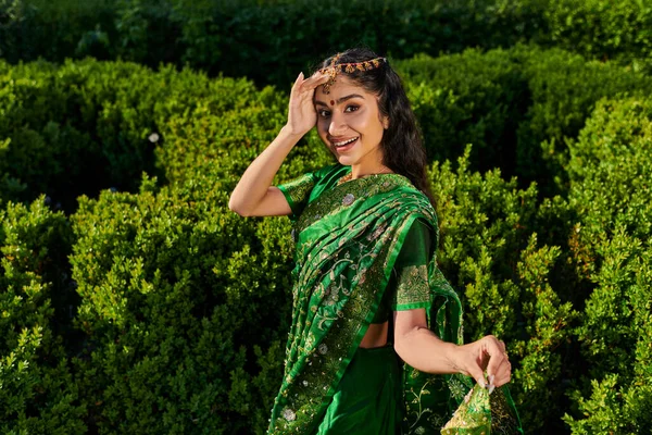 Excited and stylish indian woman in sari with pattern and matha patti near green plants outdoors — Stock Photo