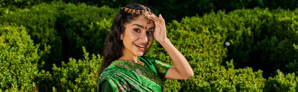 Mulher indiana animado em sari elegante e bindi olhando para a câmera perto de plantas verdes no parque, banner — Fotografia de Stock