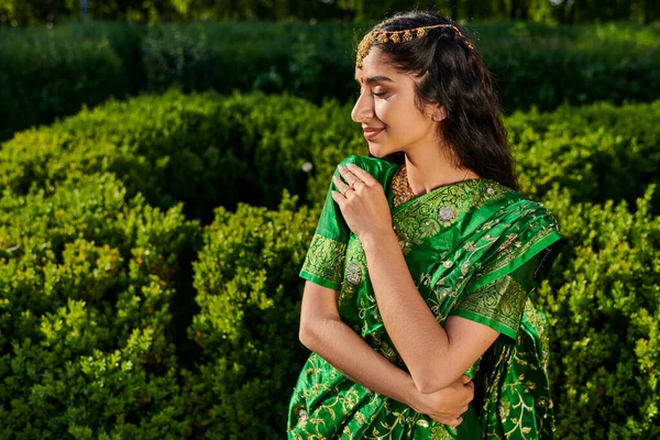 Vista lateral de mujer india joven y complacida en sari verde posando mientras está de pie cerca de plantas al aire libre - foto de stock