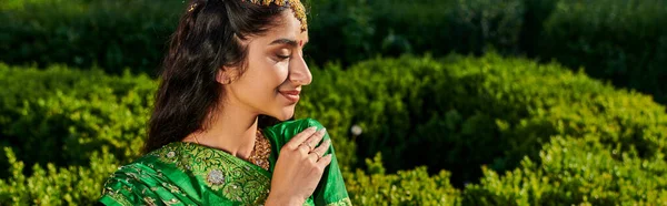 Side view of elegant young indian woman in green sari standing near blurred plants in park, banner — Stock Photo