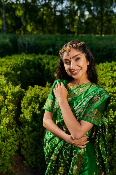 Portrait of cheerful young indian woman posing at camera in green sari in summer park — Stock Photo