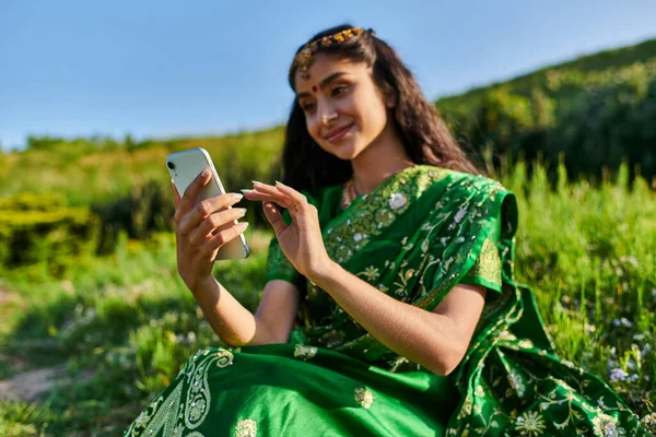 Donna indiana sorridente ed elegante in sari utilizzando smartphone mentre seduto nel verde del parco estivo — Foto stock