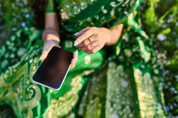 Vista cortada de jovem mulher em sari verde segurando smartphone no gramado no verão — Fotografia de Stock