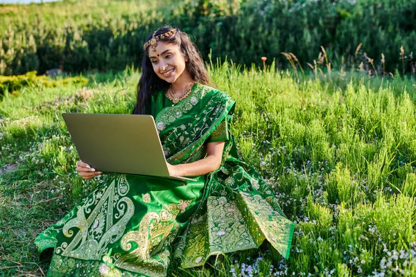 Élégant et souriant jeune femme indienne en sari à l'aide d'un ordinateur portable tout en étant assis sur la prairie en été — Photo de stock