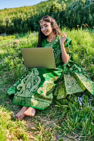 Стильная индийская женщина в сари с видеозвонком на ноутбук, сидя на лугу летом — стоковое фото
