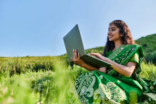 Sonriente joven india mujer en sari verde usando portátil en prado herboso en verano - foto de stock