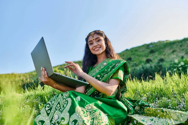 Mujer india joven positiva en sari mirando a la cámara y sosteniendo el ordenador portátil en el campo herboso - foto de stock