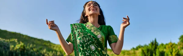 Счастливая молодая индийская женщина в зеленом сари стоит с летним пейзажем на заднем плане, баннер — стоковое фото