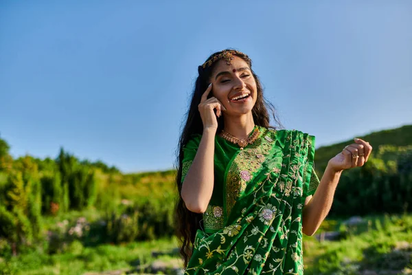 Cheerful young indian woman touching face while posing in sari with landscape on background — Stock Photo