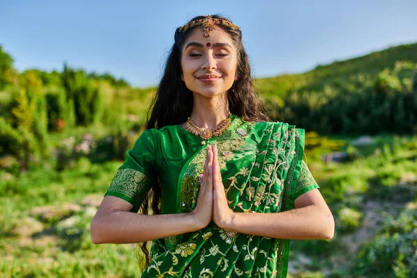 Pleased young indian woman in green sari doing praying hands gesture with landscape on background — Stock Photo