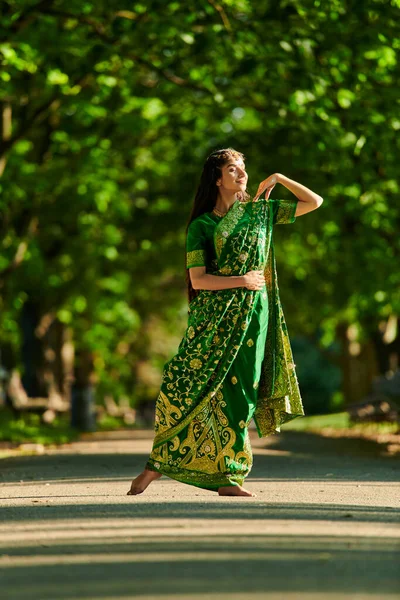 Полная длина стильная и улыбающаяся индийская женщина в сари позирует на дороге с деревьями на заднем плане — стоковое фото