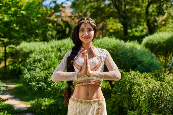 Smiling and stylish indian woman in matha patti doing praying hands gesture in park — Stock Photo