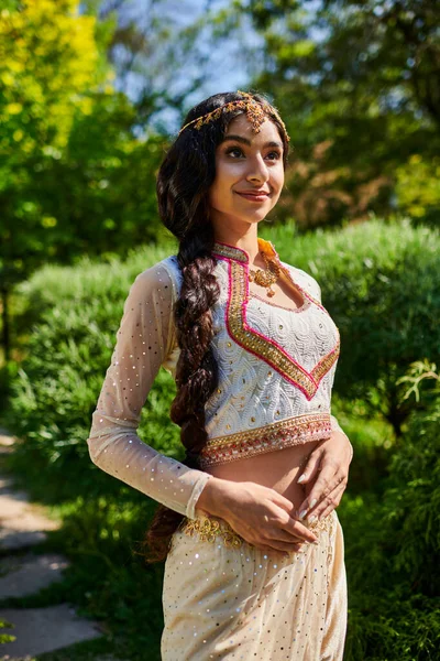 Stylish and young indian woman in trendy traditional outfit standing and posing in park — Stock Photo