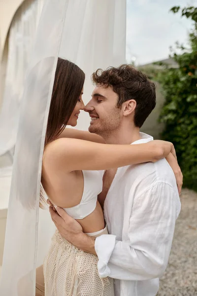 Sexy couple, happy man hugging beautiful woman while standing near white tulle of pavilion on beach — Stock Photo