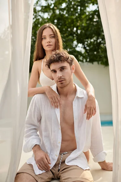 Beautiful woman embracing boyfriend with tattoo sitting in white shirt inside of beach pavilion — Stock Photo