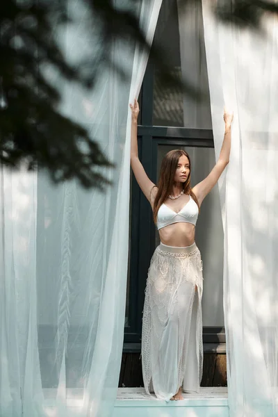 Femme gracieuse en haut de bikini et jupe posant près du tulle blanc de la maison de plage et regardant loin — Photo de stock