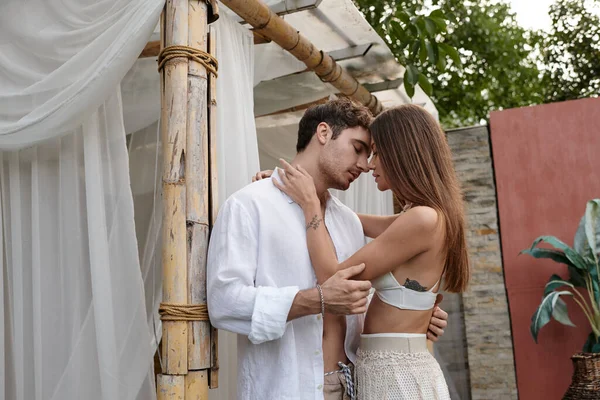 Sensual woman in white attire embracing handsome man near beach pavilion during vacation, intimate — Stock Photo