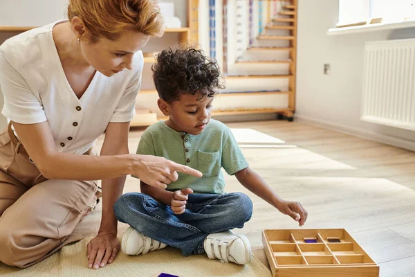 École Montessori, éducation préscolaire, professeur pointant vers une boîte en bois près d'un garçon afro-américain — Photo de stock