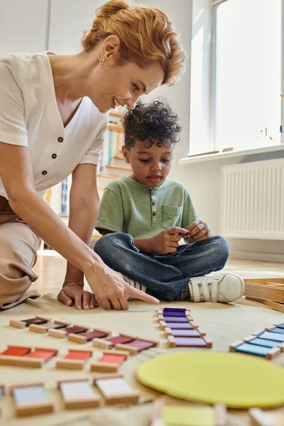 École Montessori, professeur heureux pointant vers les blocs colorés en bois près du garçon afro-américain, intelligent — Photo de stock