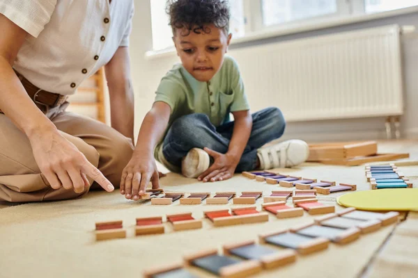 Montessori material, menino americano africano inteligente jogando jogo educativo perto do professor, colorido — Fotografia de Stock