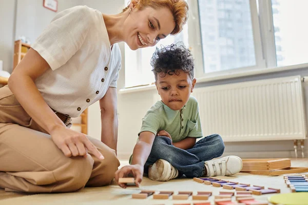 Material montessori, chico afroamericano inteligente jugando juego de color educativo cerca de maestro feliz - foto de stock