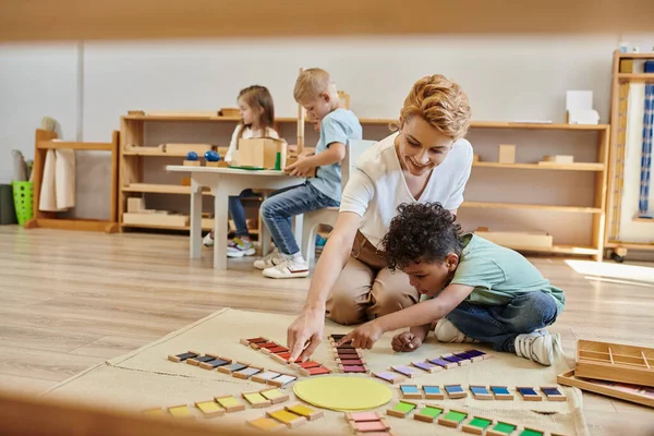 Montessori-Material, afrikanisch-amerikanischer Junge spielt pädagogisches Farbspiel in der Nähe glücklicher Lehrer, Kinder — Stockfoto
