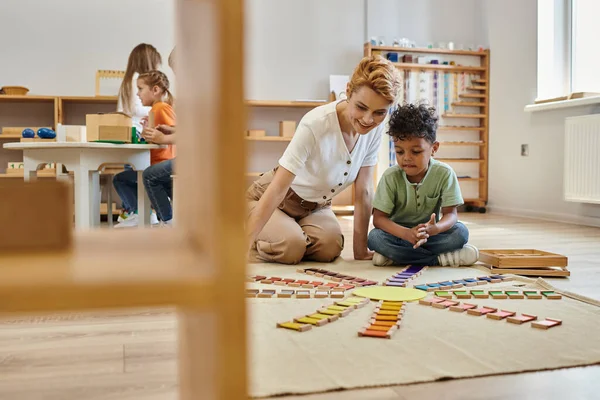 Montessori material, menino americano africano jogando jogo de cor educacional perto de professor alegre, crianças — Fotografia de Stock