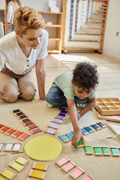 Material montessori, chico afroamericano inteligente jugando juego de color educativo cerca de la mujer feliz - foto de stock