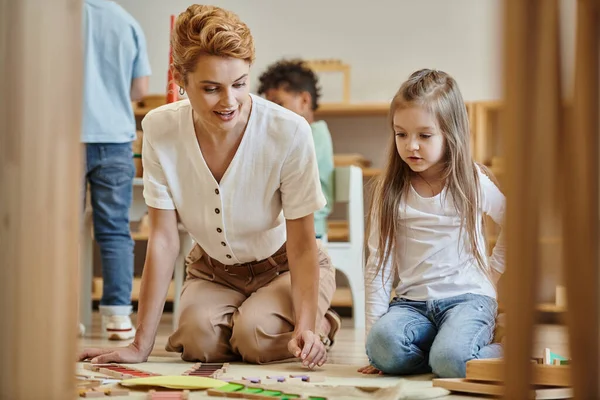 Maestra rubia y niña preescolar inteligente jugando juego montessori, mujer y niños, educativo - foto de stock