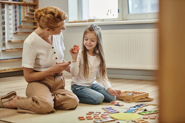 Montessori escola, menina feliz sentado perto do professor feminino segurando pedaço de madeira do jogo educativo — Fotografia de Stock