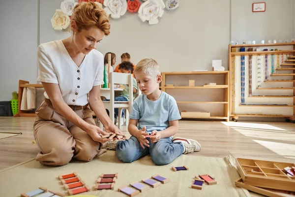 Montessori escuela, profesora sentada cerca de chico rubio y mostrando juguetes de madera, juego educativo - foto de stock