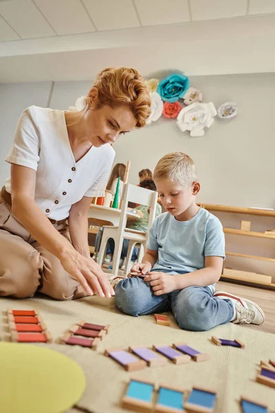 Montessori escuela, profesora sentada cerca de chico rubio jugando con juguetes de madera, juego educativo - foto de stock