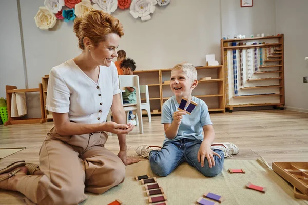 Escuela montessori, niño feliz jugando juego de combinación de colores cerca de la maestra, sentado en el suelo - foto de stock