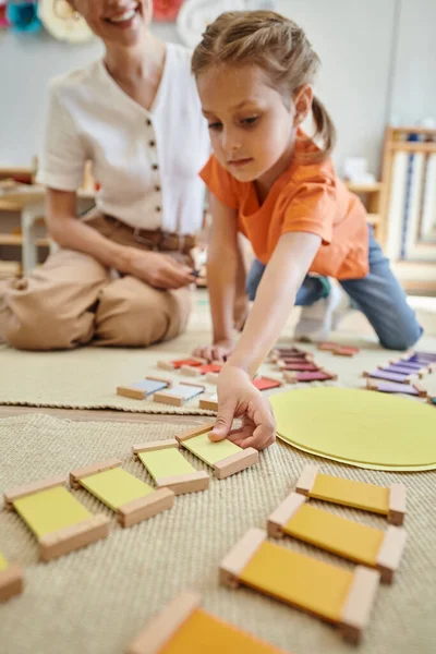 Matériel montessori, fille mignonne jouant jeu de correspondance des couleurs près de professeur femelle, assis sur le sol — Photo de stock