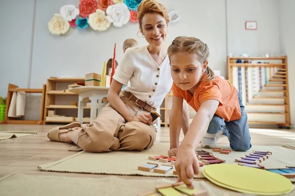 Montessori material, menina jogando jogo de correspondência de cores perto alegre professor feminino, sentado no chão — Fotografia de Stock