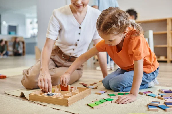 Montessori concepto de la escuela, chica jugando juego de combinación de colores cerca de profesor femenino, sentado en el suelo - foto de stock
