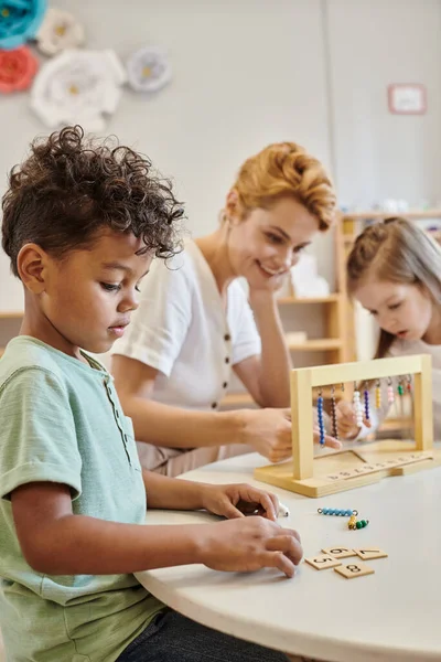 Afrikanisch-amerikanischer Junge, der Kacheln zählt und verwendet, Montessori-Konzept, Lehrer und Mädchen spielen Spiel — Stockfoto
