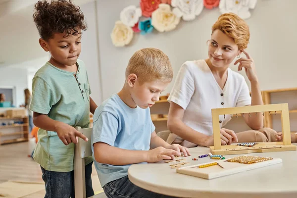 Escuela montessori, profesor feliz observando a los niños interracial, jugando juego educativo, diversos chicos - foto de stock