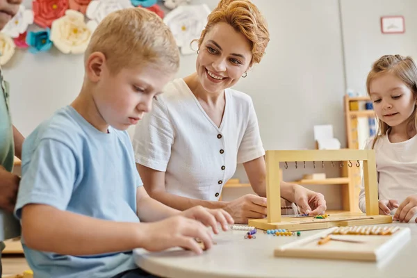 Montessori-Materialien, glückliche Lehrerin beobachtet konzentrierten Jungen in der Nähe von Kindern während des Unterrichts — Stockfoto