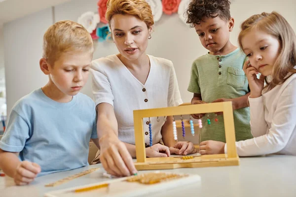 Escuela montessori, niños multiculturales jugando con escaleras de cuentas de color cerca del maestro, diversidad - foto de stock